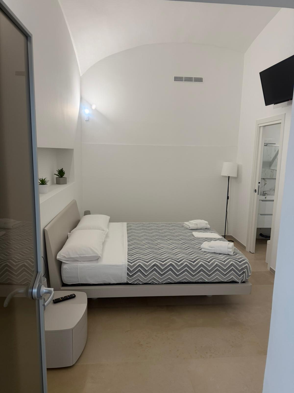 Camera moderna con letto matrimoniale, coperte piegate, luci a parete e TV montata, pareti bianche.